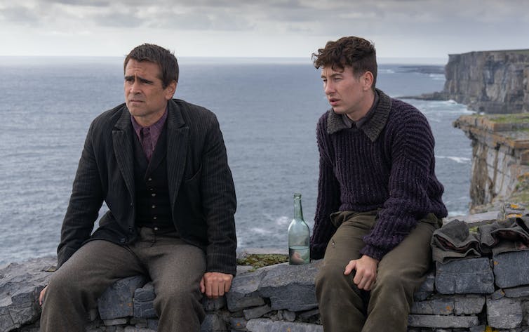 Colin Farrell and Barry Keoghan sit together on a wall overlooking the sea.