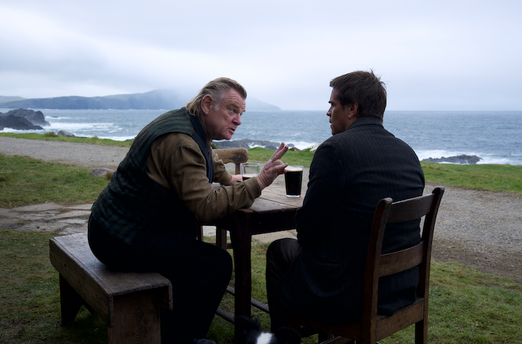 Brendan Gleeson and Colin Farrell sit at an outdoor pub table with pints, with a backdrop of the Irish coast.